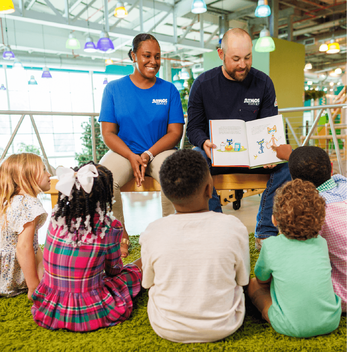 Children sitting on a rug looking at a woman and man reading a book 