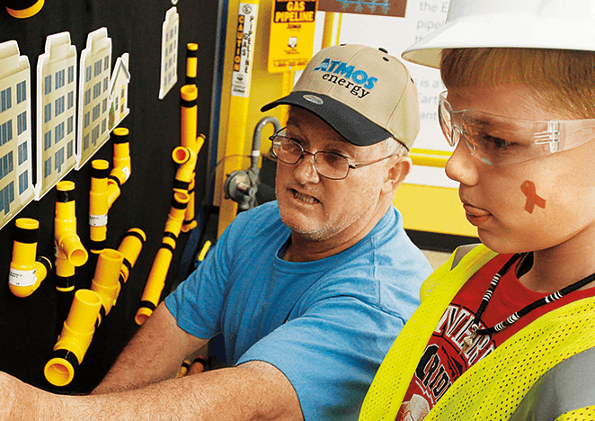 Atmos Energy employee helping a child dressed in safety gear attaching pieces of yellow pipe on a felt board that hangs on the wall.