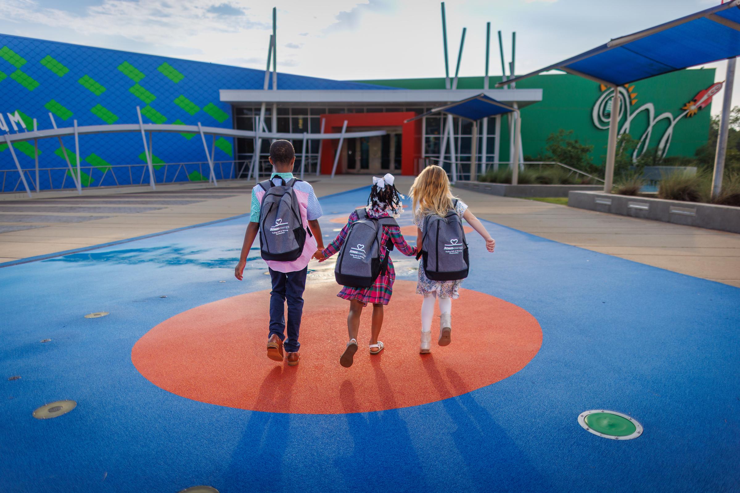 Three school aged children holding hands with gray backpacks on.