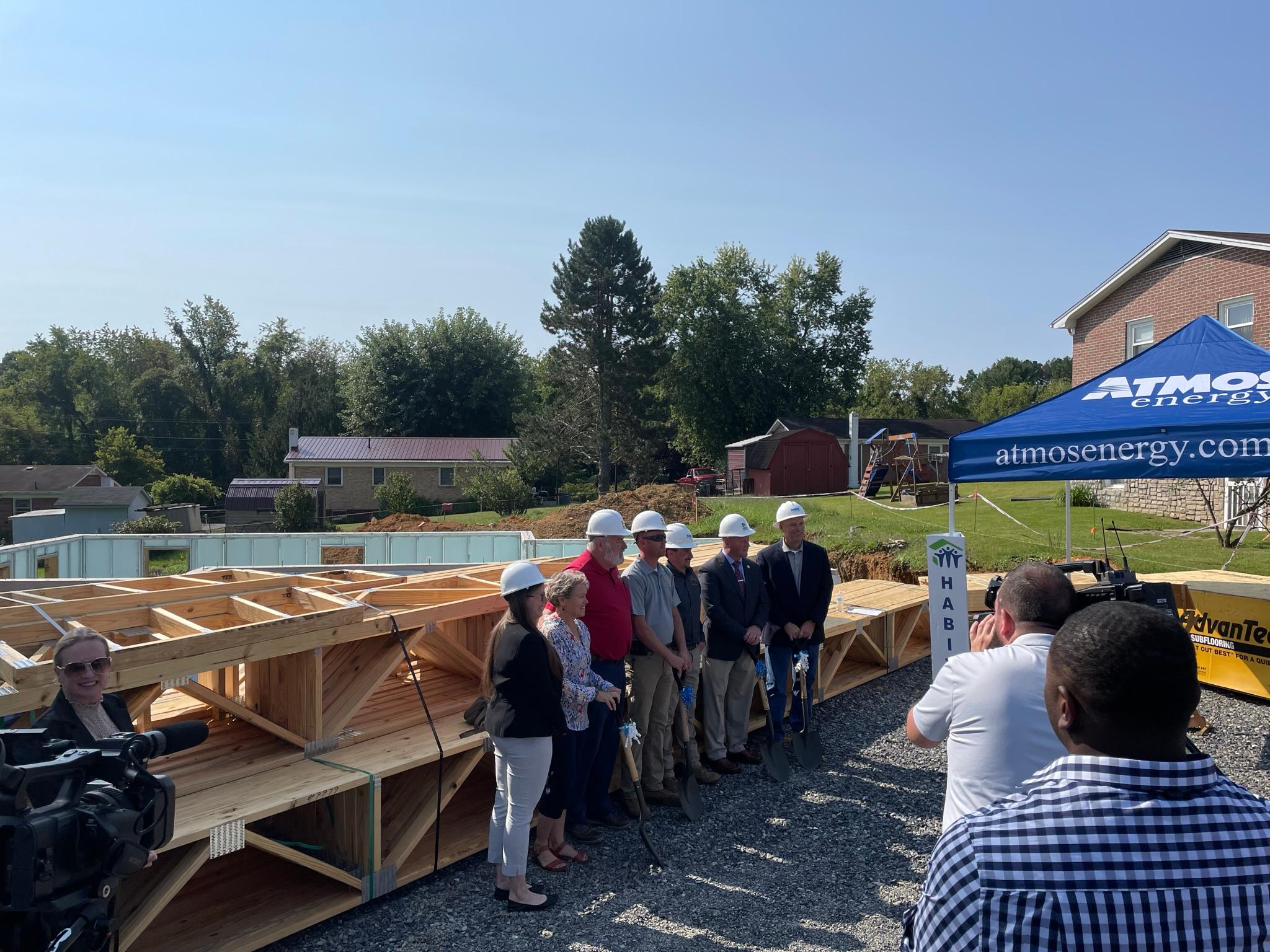 People in hard hats with shovels in front of a partially built house for a ground breaking ceremony.
