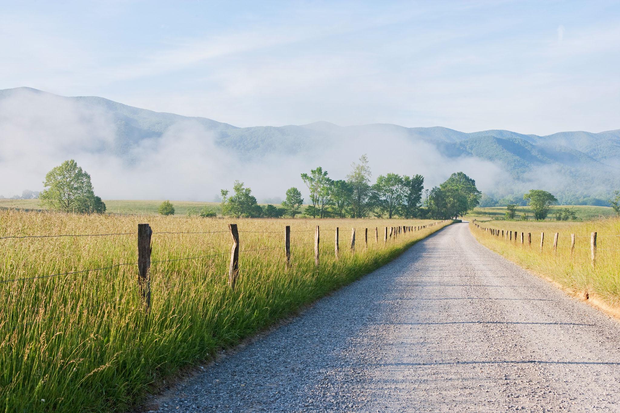 Picture of a gravel country road in Tennessee, with a wire and wood fence on either side of the road, low morning fog and a background of mountains and blue sky