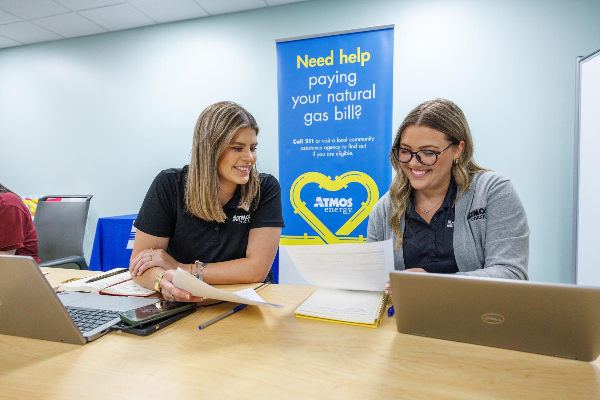 Two women smiling while sitting at a table reading some papers near laptops