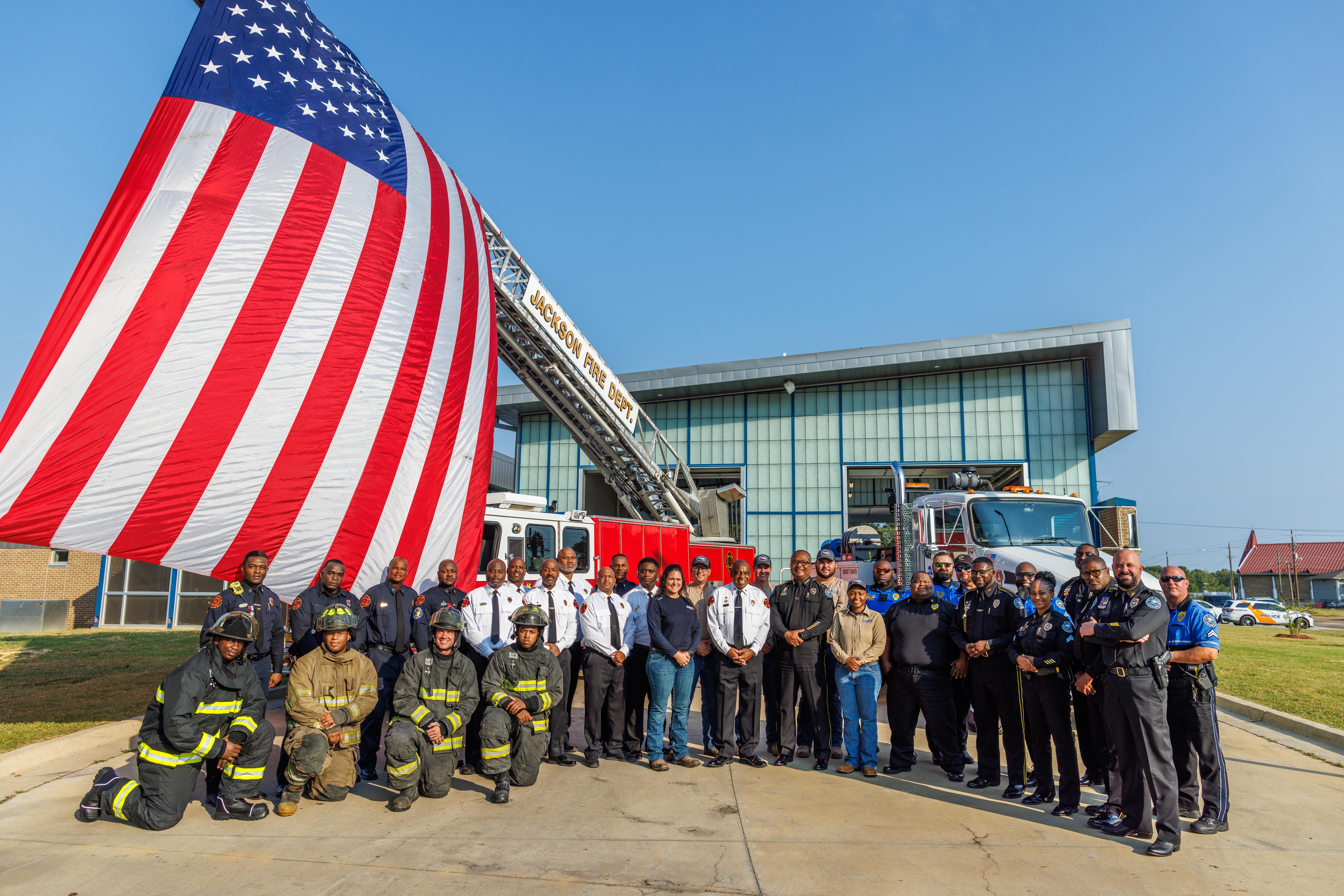 A group of men and women standing in front of a fire station with the American flag being flow from a firetruck.